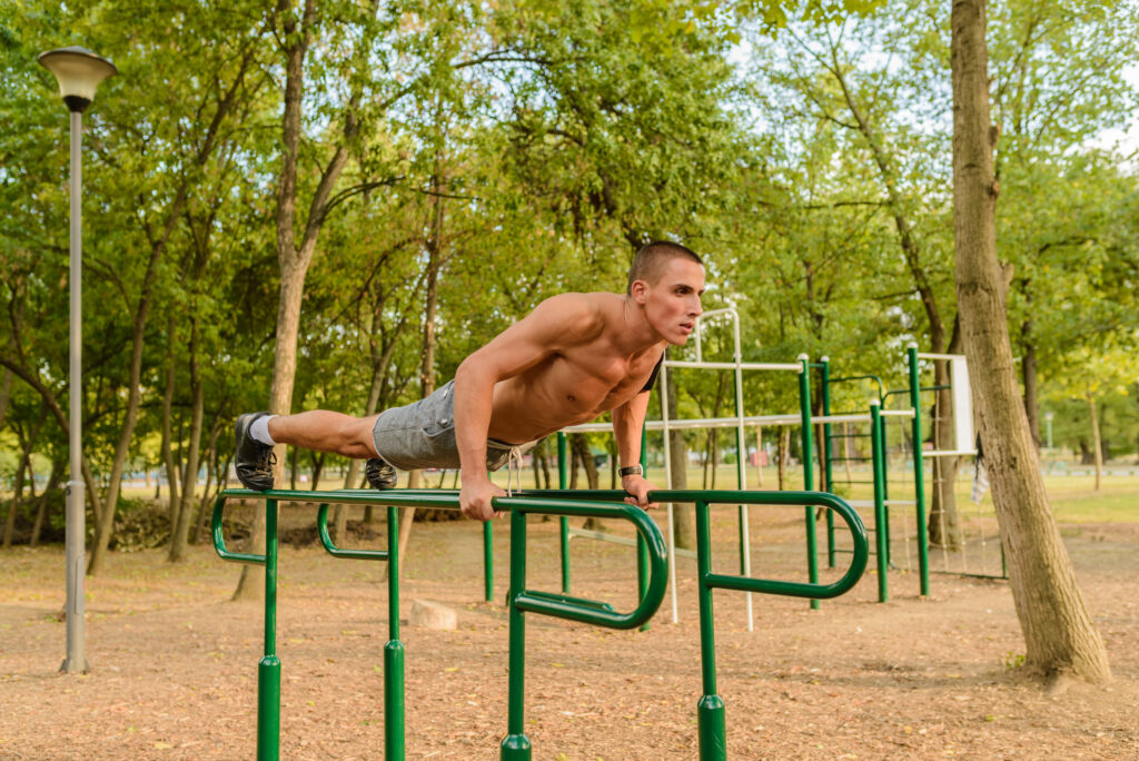 Man performing a planche exercise on parallel bars in an outdoor fitness park surrounded by trees.