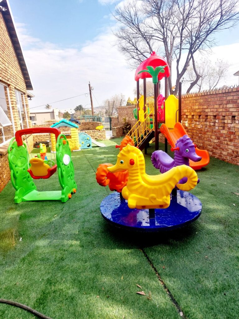 Creche play area with a carousel, seahorse spring riders, a swing and a jungle gym on artificial grass