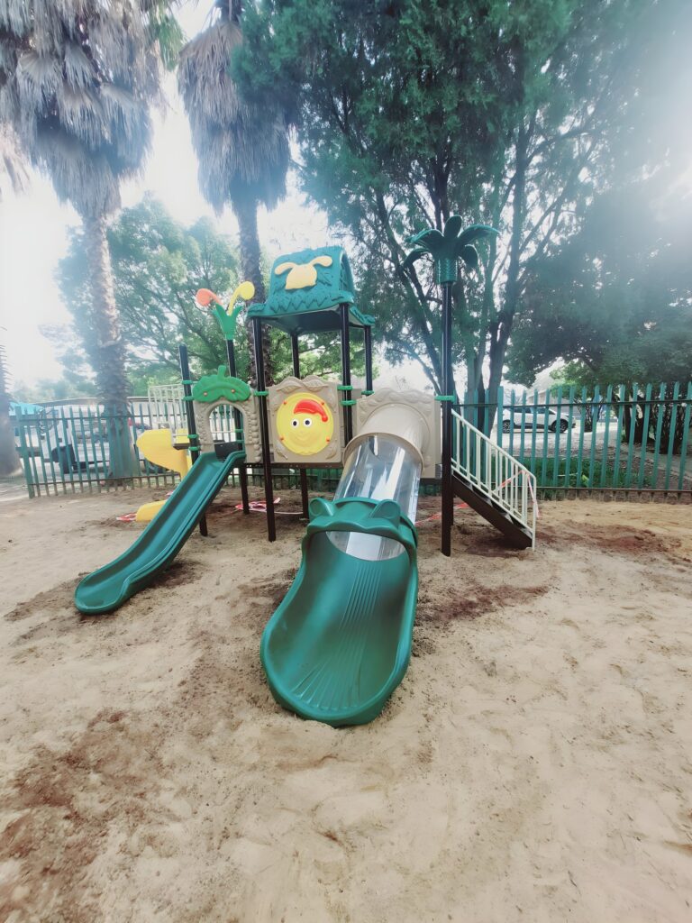 Green jungle gym with a tube slide and a wave slide in a sand play area under palm trees