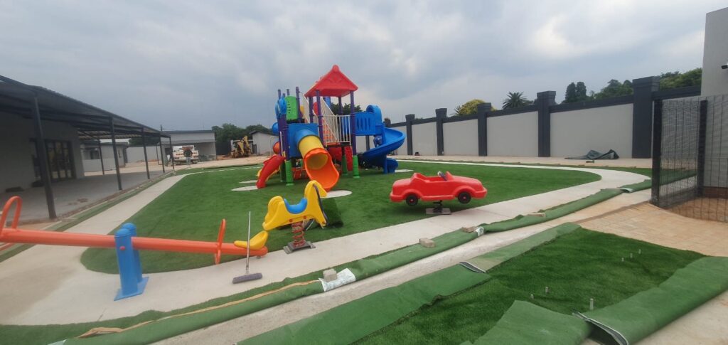 Play area with a jungle gym, a seesaw and spring riders on artificial grass beside a paved path