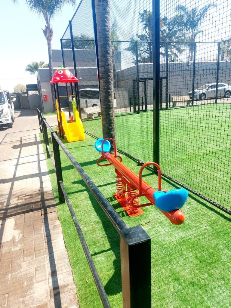 Seesaw and a small jungle gym on artificial grass beside a sports court