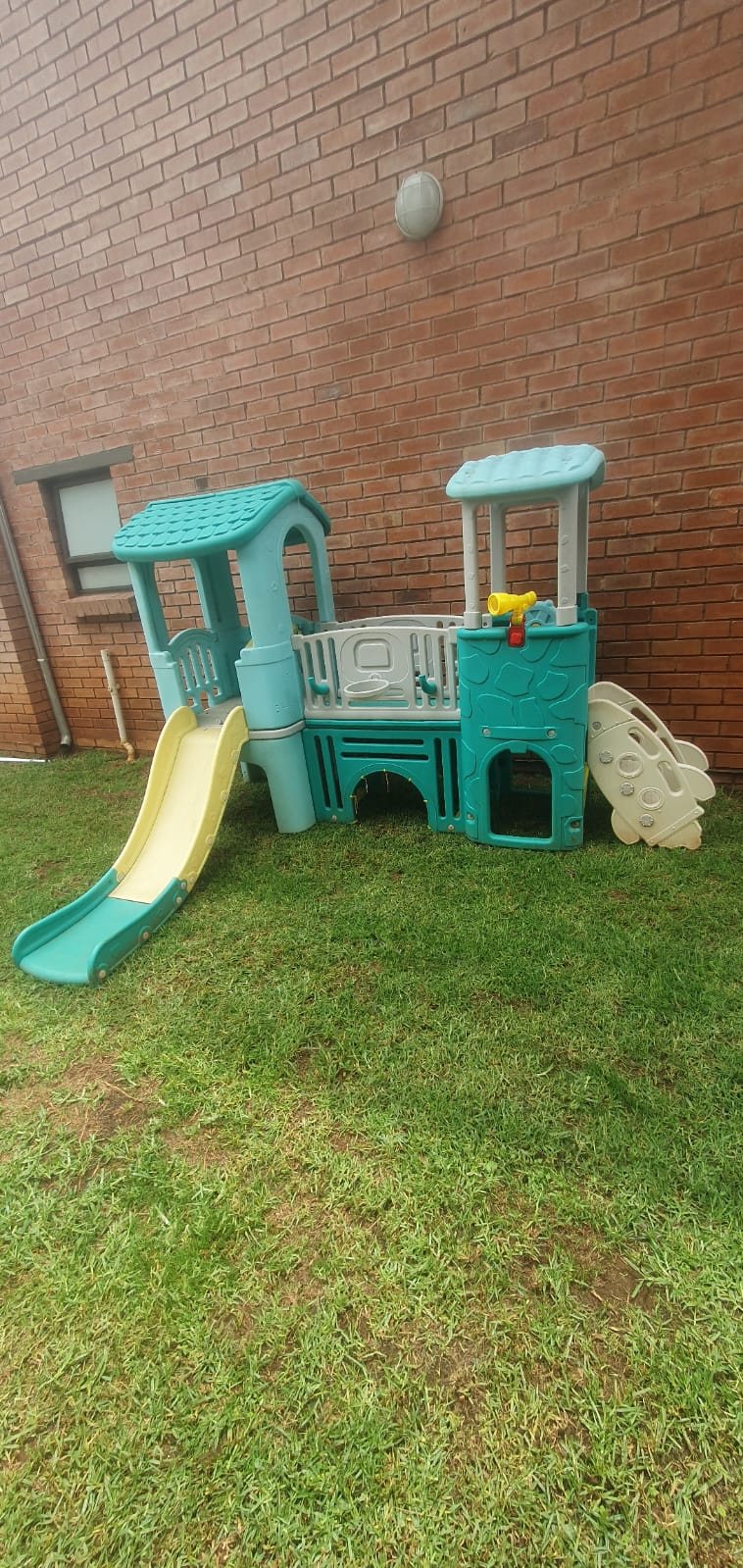Toddler playhouse with a slide on a lawn against a brick wall